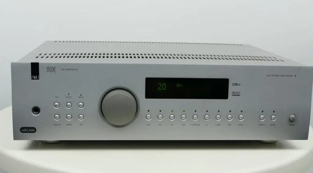 Zilveren Arcam AV8 voorversterker met duidelijk zichtbare bedieningsknoppen, digitale display en merklabels op een witte achtergrond." Bijschrift: